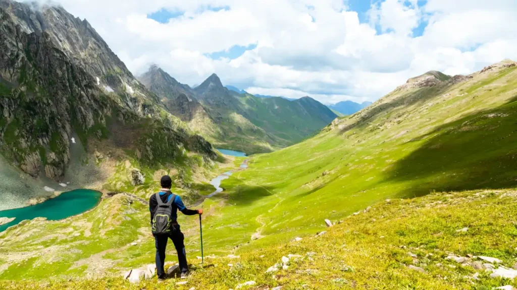 Trekker Watching a River Flow in the Lush Green Meadows with the Mountains with the Backdrop - How Difficult is the Kashmir Great Lakes Trek?